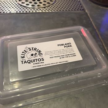 Kite String Cantina - a plastic container of taquios - A clear plastic container labeled 'Kite String Taquitos' with Poblano pork taquitos inside, placed on a metal surface near a drain. - Cocktail Bars Near Me - Chicago, Illinois