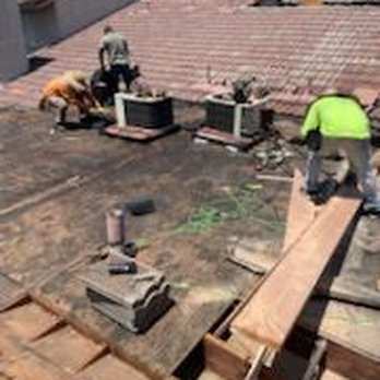 GreenCal Construction - Photo provided by Green Cal while in progress of tearing old portion of flat roof. - Three construction workers are working on a flat roof, installing or repairing roofing materials. They are using wooden planks, bricks, and tools, with some bricks placed on the roof and others being positioned by the workers. The background shows neighboring rooftops and a clear sky. - Roofing Near Me - Bellflower, California