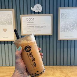 Thai Milk Tea with bobas foregrounding an interior wall that features art (defining boba) and a sign that encourages readers to "be kind."