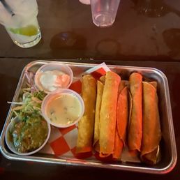 Flautas - A metal tray contains five flautas, accompanied by small cups of sour cream, guacamole, and a creamy dipping sauce. The tray is lined with checkered paper, and the food is set on a dark wooden table. In the background, there are several empty and partially filled plastic cups, some with drinks and ice, indicating a casual dining setting.