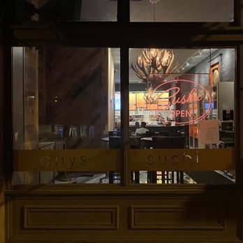 jony's sushi - The image shows a restaurant window with a neon sign reading 'Sushi OPEN' in pink, visible from outside at night. Inside, the dining area features a chandelier resembling antlers, wooden wall accents, and patrons seated at tables. The interior lighting is warm, and digital menu boards are visible behind the counter. The window has a yellow band with partially obscured text, and the exterior includes a sidewalk and building facade. - Sushi Bars Near Me - Columbus, Ohio
