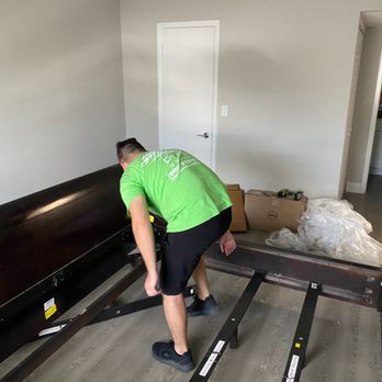 Pure Moving - A person wearing a green shirt and black shorts is assembling a bed frame in a room with light gray walls and wood flooring. The individual is bent over, working on the bed frame, which is partially assembled. There are boxes and plastic wrapping on the floor nearby, indicating ongoing furniture assembly or moving activities. - Movers Near Me - Miami, Florida