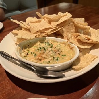 J. Alexander's Restaurant - A white round plate contains a serving of tortilla chips arranged around a small bowl of queso dip, which is garnished with chopped herbs. The plate is placed on a wooden table, and part of a person's arm and a white plate are visible in the background. - New American Near Me - Northbrook, Illinois