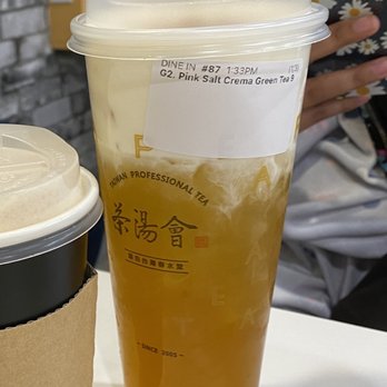 TP Tea - Pink Salt Crema Green Tea - A tall, transparent plastic cup filled with green tea topped with pink salt crema, featuring a plastic lid. The cup has a label with order details and a logo with Chinese characters, indicating it is from a Taiwanese bubble tea shop. The background shows a person holding a phone with a daisy-patterned case, seated at a table. - Bubble Tea Near Me - Richardson, Texas