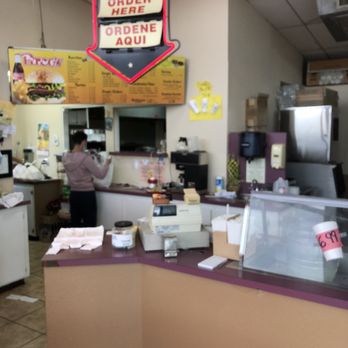 Pirco's Charbroil - Work area assembling takeaway meals - The image shows the interior of a fast-food restaurant with a counter where a person is preparing takeaway meals. A prominent illuminated sign above the counter reads 'WELCOME PIRCOS' and 'ORDER HERE' in English and Spanish. The counter has a cash register, various supplies, and a glass display case. The background includes a menu board with food options and some kitchen equipment. The setting appears to be casual and functional for quick service. - Mexican Near Me - El Paso, Texas