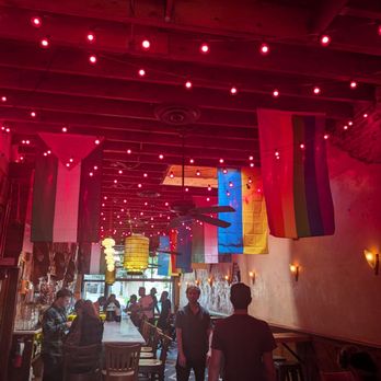 The Thin Man - The image shows the interior of a bar with a wooden ceiling and walls, decorated with rainbow flags and pink string lights hanging from the ceiling. There are several patrons standing and sitting, some at the bar and others at tables. The bar area has a counter with bar stools, and the back wall displays bottles and glasses. Warm wall-mounted lights illuminate the space, and large windows at the far end let in natural light. The overall atmosphere appears lively and festive. - Bars Near Me - Denver, Colorado
