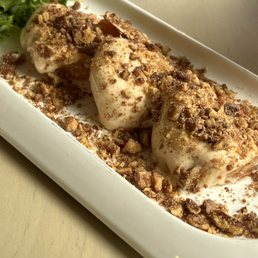 Amaretto Shrimp - Three amaretto shrimp are presented on a white rectangular plate, coated with a creamy sauce and sprinkled with crushed nuts. The dish is garnished with a small side of leafy greens, and the plate is placed on a light-colored surface. The presentation emphasizes the texture of the crushed nuts and the creamy coating of the shrimp.