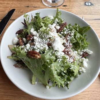 Black Cow - Pear Salad - A white ceramic bowl holds a pear salad consisting of mixed leafy greens, sliced pears, pecans, and crumbled cheese, likely feta or goat cheese. The bowl is on a wooden table, with a knife to the left and a glass of water or wine above. The salad appears fresh and garnished with cheese and nuts, typical of a dish served at a restaurant. - New American Near Me - Newburyport, Massachusetts