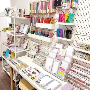 Calliope Paperie - So many things to choose from. - The image shows a stationery store display with various notebooks, pens, and decorative items. Items are organized on wall-mounted shelves, including colorful pens, markers, and notebooks with floral and pastel designs. The table in front holds additional notebooks, sticky notes, and small stationery accessories, with a stool nearby. The background features a pegboard wall with hooks for hanging supplies, and the store appears well-lit and neatly arranged. - Gift Shops Near Me - Hudson, Massachusetts