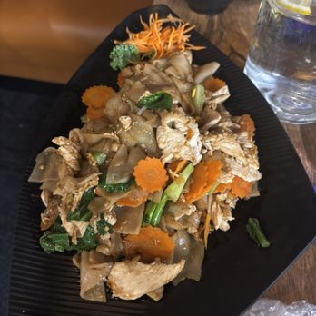 Thai Basil - Pad See U - A dish of Pad See U with vegetables, chicken, and rice is served on a black plate, accompanied by a glass of lemon water and a bowl of rice. - Thai Near Me - Madison, Wisconsin