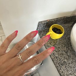 I always get simple, but they're so perfect! - The image shows a hand with manicured nails that have a pink tip design and clear bases, positioned in front of a granite countertop. The hand is wearing a silver ring with a cross on the ring finger. In the background, there is a yellow cup filled with black stones or beads, placed on the countertop, and part of a white appliance is visible.