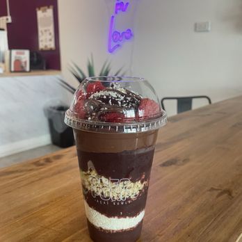 Deep Purpl - Acai Cup - A layered acai bowl in a clear plastic cup features dark acai puree, topped with strawberries, chocolate drizzle, and coconut flakes. The cup is placed on a wooden counter inside a cafe, with a neon sign reading 'People in Love' visible in the background. - Juice Bars & Smoothies Near Me - Chicago, Illinois