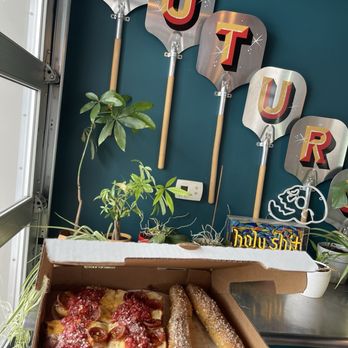 Futuro - A pizza box on a table holds a pepperoni pizza with melted cheese, two breadsticks sprinkled with salt, and a small container of dipping sauce. The background features potted plants, decorative signs spelling 'FUTUR,' and a teal wall. The setting appears to be an indoor space with a casual atmosphere, possibly a restaurant or cafe. - Pizza Near Me - Indianapolis, Indiana