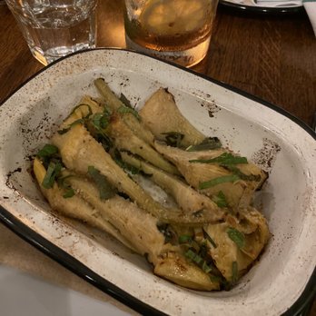 Bar Camillo - Carciofi - The image shows a white, rectangular dish containing baked artichokes garnished with chopped herbs, placed on a wooden table. Nearby, there is a glass of water and a drink with lemon slices and mint, suggesting a dining setting. The background includes a napkin and part of a plate, indicating a restaurant environment. - Italian Near Me - Brooklyn, New York