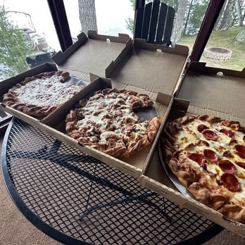 The Dock - Homemade pizza - cheese, sausage & veggie, pepperoni, pepperoni & sausage - Three large pizzas are arranged in open cardboard boxes on a round metal table. The pizzas include cheese, sausage and veggie, pepperoni, and pepperoni with sausage. The setting is indoors near a window with a view of trees, a lake, and outdoor furniture, suggesting a lakeside or cabin environment. - Pizza Near Me - Hayward, Wisconsin