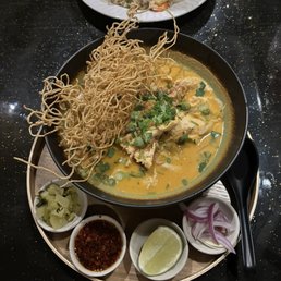Khao Soi - A bowl of Thai Khao Soi with yellow curry, chicken, green onions, and crispy noodles, accompanied by lime, pickled vegetables, chili paste, and onions.