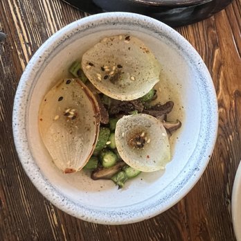 Harbinger - Morel mushroom - A ceramic bowl holds sliced onions, green vegetables, and mushrooms, seasoned with sesame seeds and spices, on a wooden table. - New American Near Me - Des Moines, Iowa