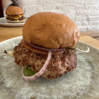 Stocked Cafe - Yummy turkey burger! - A burger with a beef patty, sliced onions, and pickles on a plate, with another burger visible in the background on a white brick wall. - New American Near Me - Brooklyn, New York