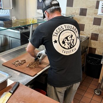 Tacos Tijuana - Preparado fresco - A man, wearing a black Tacos Tijuana taqueria T-shirt with a logo on the back and a baseball cap, is slicing cooked meat on a wooden cutting board behind a restaurant counter. The setting includes a tiled wall, a glass food shield, and kitchen equipment, indicating a food preparation area in a taqueria. - Tacos Near Me - Mesa, Arizona