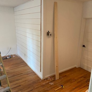 Knotty's Custom Carpentry - Work in progress. - The image depicts a corner of a room undergoing renovation, with a wooden plank leaning against the wall near a door. The room has hardwood flooring, and construction tools, including a hammer, are visible on the floor. Part of a ladder is seen on the left side, and there are electrical wires and an outlet on the wall, indicating ongoing work. The wall features a partially installed shiplap paneling on one side, with visible gaps and construction materials present. - Handyman Near Me