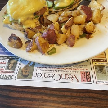 Alps Diner - Chorizo Benedict - A plate featuring two eggs Benedict with melted cheese on top, served with roasted potatoes that include chunks of potato and green pepper, on a white dish. The plate is placed on a table with a newspaper underneath, indicating a casual dining environment. - Diners Near Me - Wayne, New Jersey