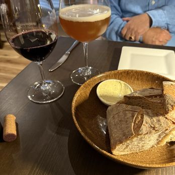 Alsace - Bread service - A restaurant table setting with a loaf of bread, a dish of butter, and two glasses of wine—red and white—alongside a person in a blue shirt. - French Near Me - Union, Maine