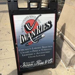 We just stumbled upon this cigar shop and it was a real treat! - A sidewalk sign advertising Iwan Ries, a tobacconist in Chicago, featuring the shop's logo, history, and product offerings like cigars, pipes, and accessories.