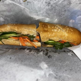 Wrap N Roll - Beef 10. Lemongrass Beef Sandwich - A Vietnamese banh mi sandwich is shown, cut in half and placed on white paper. The sandwich contains fresh vegetables such as cucumber, carrots, and cilantro, along with slices of meat inside a baguette-style bread. The bread has a golden-brown crust with sesame seeds on top. The filling is visible, with vibrant colors of the vegetables contrasting with the bread. - Vietnamese Near Me - Albuquerque, New Mexico