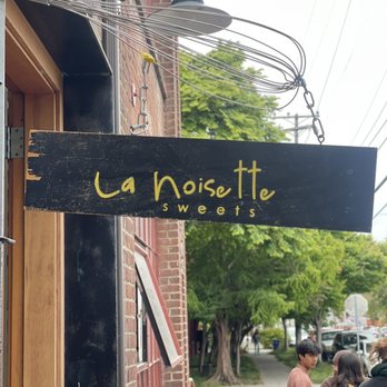 La Noisette Sweets - A black wooden sign with yellow handwritten-style lettering displays 'La Noisette Sweets' and is mounted outside a bakery on a brick building. The sign is attached with metal chains and is positioned near a sidewalk with pedestrians and trees in the background. Overhead, electrical wires and a street lamp are visible, indicating an urban setting. - Bakeries Near Me - Berkeley, California