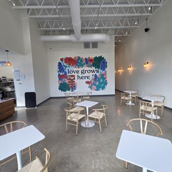 Bagel Bear - Great mural! - The image shows the interior of a cafe with a high ceiling and exposed ductwork. A large mural on the back wall displays vibrant floral artwork and the phrase 'love grows here.' The space has a polished concrete floor, white walls, and minimalistic lighting fixtures. Multiple white tables with wooden chairs are arranged throughout the area, providing seating for customers. The overall atmosphere is modern and clean, with a focus on welcoming decor. - Bagels Near Me - Cedar Park, Texas
