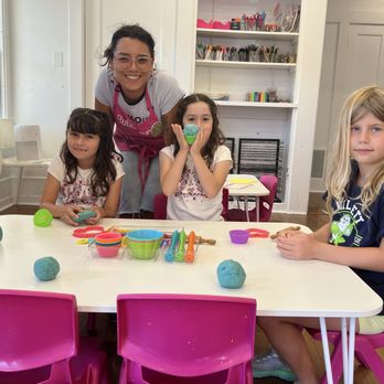 Bubblegum Art Studio - Elle is the best! - A woman and three young girls are seated at a white table in a classroom, surrounded by colorful arts and crafts supplies such as playdough, cookie cutters, and utensils. The woman is smiling and leaning over the table, while the girls are engaged with the materials. The classroom has white walls, a window with natural light, and shelves with various art supplies in the background. - Art Classes Near Me - Davis, California
