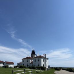 A lighthouse and adjacent buildings are located near the shoreline, with a clear blue sky overhead and a grassy area in the foreground.