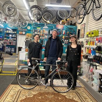 Broadway Bikes - One of the first customers at the new Broadway Bikes - Inside a bicycle shop, three people stand together, smiling at the camera. The shop has bikes hanging from the ceiling and various cycling accessories displayed on the walls and shelves. The group includes two women and one man, with the women wearing black overalls and the man dressed casually with a beanie. A bicycle is positioned in front of them on a rug, and the shop's interior features organized tools and cycling gear, indicating a well-stocked bike repair and sales store. - Bikes Near Me - Chicago, Illinois