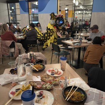Upside on Moore - The image shows a lion dance costume performing in a food hall with multiple tables and diners. The food hall has modern decor with large windows, lighting fixtures, and a mix of seated diners and standing patrons. Food and drinks are visible on the tables, including bowls of noodles, plates with various dishes, and beverages. The lion costume is yellow with black and red details, and the performer is in mid-movement among the seated diners. - Food Court Near Me - Arlington, Virginia
