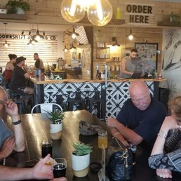 :Downshift Brewing Co - The image shows a group of people seated around a dark wooden table inside a brewery or restaurant. There are drinks, including glasses of red wine and other beverages, on the table along with potted plants as decoration. In the background, a bar area with a bartender working on a laptop is visible, with a wooden wall and shelves holding various items. A large light fixture with exposed bulbs hangs above the table, and a sign reading 'ORDER HERE' is mounted on the wall behind the bar.