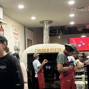 Timber Pizza - McLean - Wood  fired oven! - Employees in red aprons and caps work around a wood-fired pizza oven at Timber Pizza in McLean, VA, with a white brick wall and a digital screen in the background. - Pizza Near Me - McLean, Virginia