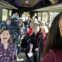 First stop on bus - A group of women is seated inside a bus, celebrating a 55th birthday. The bus interior has decorations such as balloons and banners, with some women wearing sunglasses and hats. They are smiling, laughing, and appear to be enjoying the celebration. The background shows windows with a view of trees outside, and a woman is standing at the back, possibly taking photos or preparing for the event.