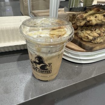 Ape Coffee - Monkey Business - A plastic cup of iced coffee with whipped cream and banana slices, featuring the Ape Coffee logo, placed on a counter near pastries and drinks. - Cafes Near Me - Placentia, California