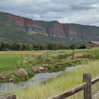 James Ranch Grill - Views - The image depicts a rural landscape with lush green fields and a small stream running through the foreground. A wooden fence runs parallel to the stream, and there is a barn or shed on the right side. In the background, layered red and green mountains rise under a cloudy sky, creating a natural and peaceful setting. - New American Near Me - Durango, Colorado