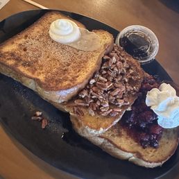 French Toast - A black oval plate holds two slices of French toast topped with whipped cream, chopped nuts, and syrup. The toast appears golden brown and is garnished with a dollop of whipped cream and a small container of syrup. The setting includes a wooden table with other dishes and utensils visible in the background.