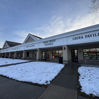 Third Proof Pizzeria - The image shows a shopping plaza with several storefronts, including Third Proof Pizzeria, with a clear sky overhead. Snow covers the ground in front of the stores, and a sidewalk runs parallel to the storefronts. A person is seen entering the pizzeria, and the storefront has large windows with an illuminated 'Open' sign visible inside. - Pizza Near Me - Pine Brook, New Jersey