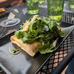 A dish featuring greens and cheese on a black rectangular plate is placed on an outdoor metal table with green glasses in the background.