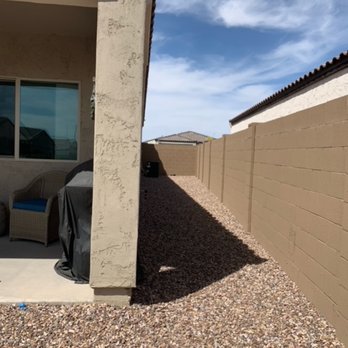 Diamond Cut Landscaping - side of backyard before - The image shows a narrow side yard of a residential property with gravel ground covering. On the left, part of the house's exterior wall and a patio area with outdoor furniture and a covered grill are visible. To the right, a tall beige fence runs along the length of the yard, casting a shadow on the gravel. The sky above is partly cloudy with blue sky visible, and neighboring rooftops can be seen in the background. - Landscaping Near Me - Phoenix, Arizona