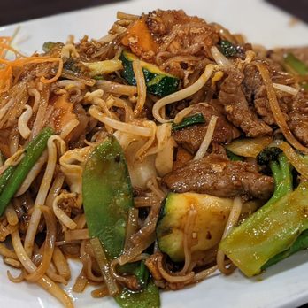 Chilli Thai - Paid Thai Beef with extra vegetables. - A rectangular white plate holds a serving of Thai beef stir-fry with sliced beef, broccoli, zucchini, bean sprouts, and other vegetables coated in a brown sauce. Shredded carrots are placed on the side of the plate. A metal spoon is on the right side of the plate, and the dish appears to be served in a restaurant setting. - Thai Near Me - Carrollton, Texas