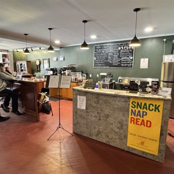 Book Bar - The image shows the interior of a cafe with a concrete counter, a chalkboard menu on the wall, and a seating area with customers. The space has warm lighting from pendant lamps, wooden flooring, and a mix of furniture including bar stools and chairs. There are coffee machines and various supplies on the counter, and a large refrigerator is visible on the right. A sign on the counter reads "SNACK NAP READ." The overall setting appears inviting and suitable for casual dining or coffee breaks. - Coffee & Tea Near Me - Palmyra, Pennsylvania