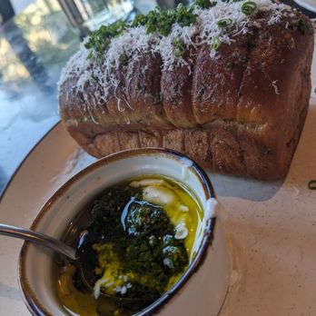 Geppetto - Pull apart soft garlic bread with pesto butter yogurt dip thing - A large, toasted loaf of garlic bread is topped with grated cheese and chopped green herbs, placed on a plate. In front of the bread, there is a small bowl containing a mixture of herbs and oil, likely for dipping. The setting appears to be a restaurant with a window view and tableware visible in the background. - Italian Near Me - Cambridge, Massachusetts