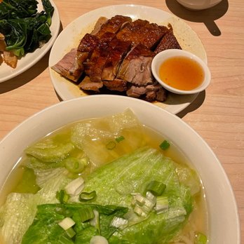 Congee Queen - Bbq duck rice noodles - A bowl of noodle soup contains broth, large lettuce leaves, and chopped green onions. A plate of roasted meat with crispy skin and sliced portions is accompanied by a small bowl of dipping sauce. The dishes are placed on a light wooden table, with an empty bowl and a spoon visible in the background. - Chinese Near Me - Markham, Ontario
