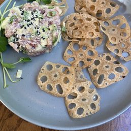Tuna Tartare - A gray ceramic plate holds a serving of tuna tartare, garnished with chopped green onions and black sesame seeds. The tartare is accompanied by several slices of crispy lotus root chips, arranged around the tartare. A few sprigs of microgreens are placed on the side, adding a fresh element to the presentation. The dish is set on a wooden table, highlighting a modern American eatery style with a focus on fresh ingredients.