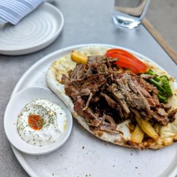 Gyro - A lamb gyro featuring sliced cooked lamb meat, tomato slices, and cucumber on a pita bread, served on a white plate. A small bowl of yogurt sauce garnished with herbs is placed beside the gyro. The meal is set on a gray table with a glass of water nearby, indicating a casual dining setting.