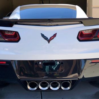 Windows & Wheels Auto Detailing - A white Chevrolet Corvette sports car is parked in a driveway, showing its rear end. The car features black accents on the rear spoiler and lower bumper, with distinctive taillights and a Corvette emblem in the center. Four exhaust pipes are visible at the bottom, and the vehicle is positioned in front of a garage door with a concrete surface beneath it. - Auto Detailing Near Me - Gilbert, Arizona